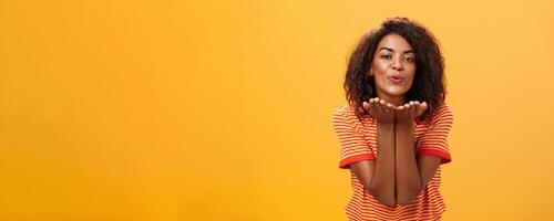 enviando apaixonado beijo para a maioria amoroso pessoa. romântico atraente e à moda jovem africano americano namorada com encaracolado Penteado dobrar para Câmera com leve sorriso, guardada lábios sopro mwah foto