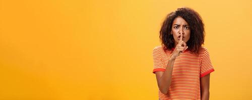 shh Faz não perturbar. retrato do sério mandão e descontente fofa africano americano menina com afro Penteado franzindo a testa calar às Câmera com índice dedo sobre boca proibindo falar alto foto