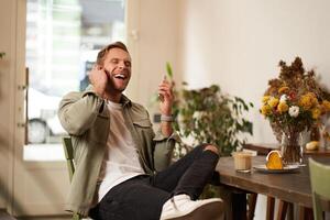 estilo de vida retrato do bonito jovem homem, sentado dentro cafeteria, falando para uma amigo em Móvel telefone com sem fio fones de ouvido, rindo e sorridente, ouvindo para engraçado podcast foto