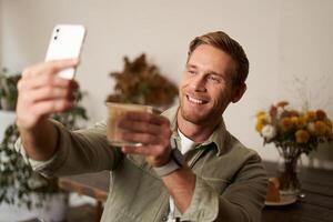 retrato do bonito sorridente loiro homem, levantando vidro do café, chats em Smartphone aplicativo, levando selfie com cappuccino dentro cafeteria, olhando feliz foto