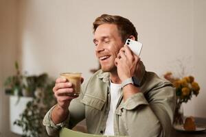 retrato do jovem bonito homem falando em a telefone, bebidas café e ri. uma cara senta dentro cafeteria tendo uma legal, agradável conversação sobre copo do cappuccino foto