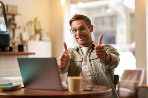 alegre sorridente cara dentro copos, homem mostra polegares acima, senta dentro cafeteria com computador portátil, recomenda local na rede Internet, e-learning serviço ou co-trabalho espaço foto