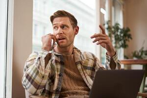 retrato do sério homem de negocios falando sobre a telefone, sentado dentro cadeira dentro uma cafeteria com computador portátil, trabalhando em projeto, discutindo relacionado ao trabalho problemas em Telefone, parece atrás ele foto