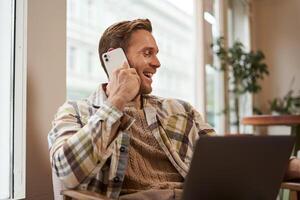 imagem do feliz cafeteria Visitante, homem com computador portátil, chamando uma amigo sobre a telefone. homem de negocios falando em Telefone, trabalhando conectados a partir de café comprar, rindo e sorridente foto