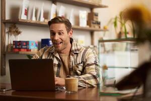 retrato do bonito jovem digital nômade, homem trabalhando dentro cafeteria em computador portátil, olhando feliz e satisfeito com dele conectados projeto, sentado dentro co-trabalho café fazer compras foto