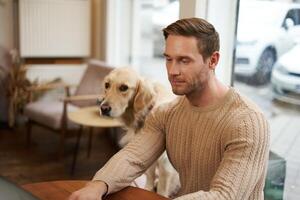 bonito jovem homem, digital nômade sentado com dele cachorro dentro uma aceita animais de estimação cafeteria, trabalhando em computador portátil enquanto uma dourado retriever relógios ele foto