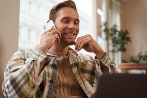 bonito homem de negocios dentro café comprar, sentado dentro cafeteria com computador portátil e chamando alguém, homem falando em a telefone com cliente, fazer inquérito sobre projeto, falando para colega de trabalho enquanto trabalhando remotamente foto