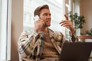 imagem do homem sentindo-me estressado enquanto falando sobre a telefone. homem de negocios sentado dentro cafeteria com computador portátil, respondendo uma chamar, ouvir mau notícia sobre a Telefone foto