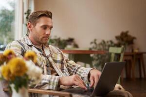 estilo de vida e local de trabalho conceito. jovem bonito homem dentro casual roupas, sentado dentro cafeteria com computador portátil, estudando on-line, trabalhando remotamente a partir de café fazer compras foto