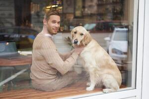 ao ar livre tiro do uma aceita animais de estimação cafeteria Onde homem senta perto a janela com dele fofo dourado retriever e sorrisos às Câmera. café fazer compras Visitante com uma cachorro foto