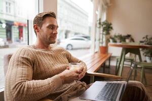 retrato do masculino empreendedor, digital nômade sentado dentro cafeteria com vidro do capuccino, olhando às computador portátil, assistindo , atendendo encontro conectados a partir de café fazer compras foto