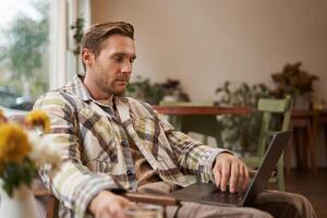 estilo de vida e local de trabalho conceito. jovem bonito homem dentro casual roupas, sentado dentro cafeteria com computador portátil, estudando on-line, trabalhando remotamente a partir de café fazer compras foto