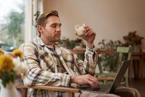 bonito loiro homem com computador portátil, sentado dentro cafeteria e bebendo café, trabalhando on-line, freelancer, conjunto acima área de trabalho lado de fora do a escritório meio Ambiente foto