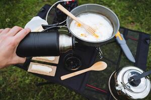 uma turista derrama quente chá a partir de uma garrafa térmica para dentro uma caneca, frito mexidos ovos dentro uma fritar panela em uma caminhada, café da manhã dentro natureza, turista comida, caminhada cozinha, chaleira colher foto