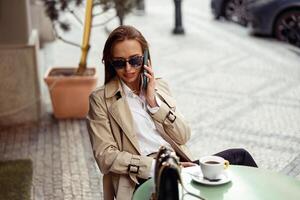 à moda mulher vestindo oculos de sol sentado às cafeteria terraço falando em telefone enquanto bebendo café foto