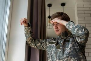 sério cabelo curto jovem homem dentro militares uniforme soldado olhando através a janela, chateado soldado sofrimento a partir de pós traumático síndrome depois de retornando lar, cópia de espaço, fechar-se. foto