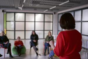 uma mulher faz uma discurso em estágio, colegas ouço para dela discurso e sentar em cadeiras e pufes dentro a corredor foto