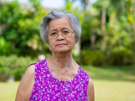 Senior mulher vestindo copos, sorridente, e olhando às a Câmera enquanto em pé dentro uma jardim. espaço para texto. conceito do envelhecido pessoas e cuidados de saúde foto