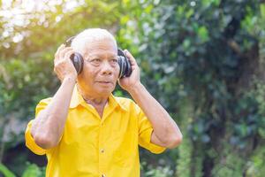 Senior homem vestindo sem fio fones de ouvido ouvindo para uma favorito música e olhando acima enquanto em pé dentro uma jardim. espaço para texto. conceito do envelhecido pessoas e relaxamento foto