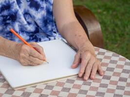 mãos do uma Senior mulher escrevendo uma livro enquanto sentado em uma cadeira dentro a jardim. fechar-se foto com cópia de espaço para texto. conceito do envelhecido pessoas e cuidados de saúde