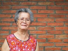 Senior mulher sorridente e olhando às a Câmera enquanto em pé dentro uma jardim com uma tijolo parede fundo. espaço para texto. conceito do envelhecido pessoas e cuidados de saúde foto