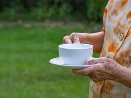 fechar-se do Senior mulher mãos segurando uma branco copo enquanto em pé dentro uma jardim com cópia de espaço para texto. conceito do envelhecido pessoas e relaxamento foto