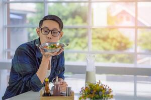 uma jovem homem segurando e olhando às a frito ovo servido em uma panela com colorida coberturas servido em uma panela enquanto sentado em uma cadeira dentro a cozinha sala. espaço para texto. simples Comida para café da manhã foto