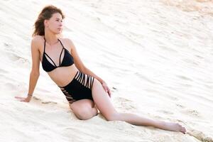 retrato do jovem mulher dentro Preto roupa de banho relaxante em arenoso de praia foto