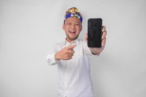 sorridente jovem balinesa homem vestindo branco camisa e tradicional cocar apontando para dele Smartphone, apresentando em branco tela cópia de espaço foto