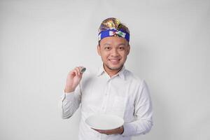 retrato do uma jovem balinesa homem dentro branco camisa e tradicional cocar mostrando e apresentando a esvaziar prato com cópia de espaço enquanto sorridente alegremente para a Câmera foto