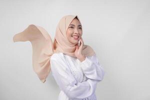elegante jovem ásia muçulmano mulher vestindo branco vestir com hijab acenando dentro a vento, modelagem pose em branco fundo foto