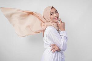 elegante jovem ásia muçulmano mulher vestindo branco vestir com hijab acenando dentro a vento, modelagem pose em branco fundo foto