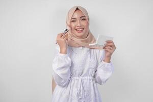 lindo jovem ásia muçulmano mulher vestindo branco vestir e hijab aplicando faço acima usando escovar. moda e cosméticos beleza conceito foto