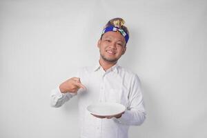 sorridente jovem balinesa homem dentro branco camisa e tradicional cocar mostrando e apresentando a esvaziar prato com cópia de espaço foto