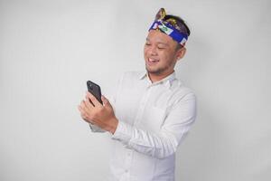 animado balinesa homem vestindo branco camisa e tradicional cocar sorridente alegremente enquanto usando Smartphone, respondendo mensagem, lendo notícia foto
