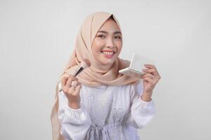 lindo jovem ásia muçulmano mulher vestindo branco vestir e hijab aplicando faço acima usando escovar. moda e cosméticos beleza conceito foto