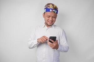 animado balinesa homem vestindo branco camisa e tradicional cocar sorridente alegremente enquanto usando Smartphone, respondendo mensagem, lendo notícia foto