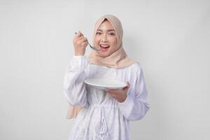com fome jovem ásia muçulmano mulher dentro hijab segurando colher e comendo a partir de a esvaziar prato com cópia de espaço sobre. Ramadã conceito foto