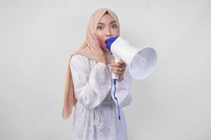 muito feliz ásia mulher vestindo hijab e branco vestir gritando para a megafone fazer anúncio, em pé sobre isolado branco fundo foto