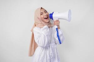 muito feliz ásia mulher vestindo hijab e branco vestir gritando para a megafone fazer anúncio, em pé sobre isolado branco fundo foto