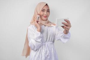 lindo jovem ásia muçulmano mulher vestindo branco vestir e hijab aplicando faço acima usando escovar. moda e cosméticos beleza conceito foto