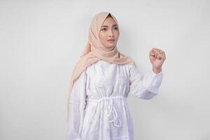 jovem ásia muçulmano mulher vestindo branco vestir e hijab com sério expressão levantando uma cerrado punho gesto sobre isolado branco fundo foto