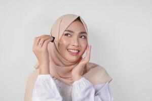 animado jovem ásia muçulmano mulher vestindo branco vestir e hijab teste ou aplicando pele Cuidado sérum em face. facial e beleza conceito foto