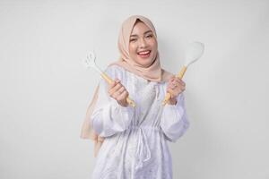 retrato do uma feliz ásia muçulmano mulher dentro véu hijab segurando espátula e cozinha cozinhando utensílios enquanto sorridente alegremente. Ramadã conceito foto