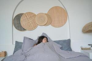 ásia mulher guardando olhos fechadas e cobertura corpo com cinzento cobertor, dormindo em cama às quarto. feriado relaxante conceito. foto