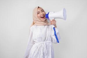 muito feliz ásia mulher vestindo hijab e branco vestir gritando para a megafone fazer anúncio, em pé sobre isolado branco fundo foto