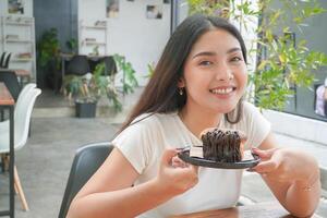 jovem ásia mulher segurando e mostrando uma Preto prato do rosquinha pastelaria nomeado cromboloni com chocolate o preenchimento para lanche tempo, sorridente com uma feliz e alegre expressão foto