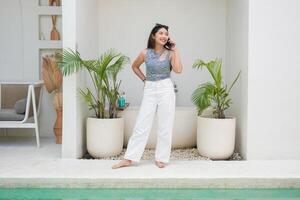 feliz ásia mulher em pé de a piscina enquanto chamando e falando em uma Móvel telefone com uma alegre sorrir em a face foto