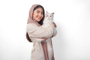 atraente jovem ásia muçulmano mulher dentro véu hijab sorridente enquanto posando e abraçando uma branco boneca de pano gato animal foto