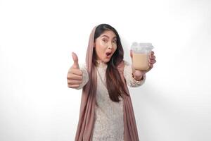 animado ásia muçulmano mulher dentro lenço de cabeça véu hijab dando polegares acima enquanto segurando café dentro uma plástico copo esperando para iftar para bebida. Ramadã conceito foto